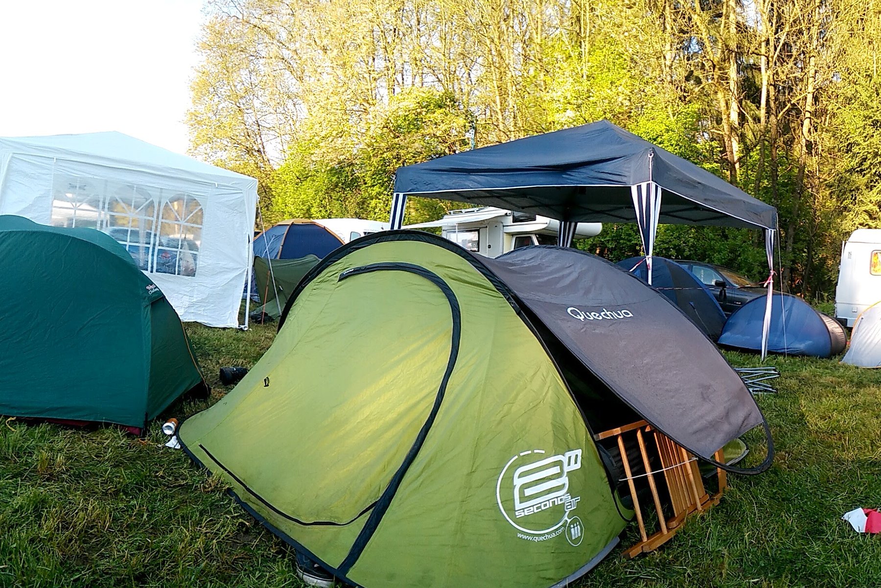 Grünes Quechua-Wurfzelt mit geöffnetem Eingang auf einer Campingwiese, daneben ein Pavillon, ein Campingstuhl und Wohnmobile.