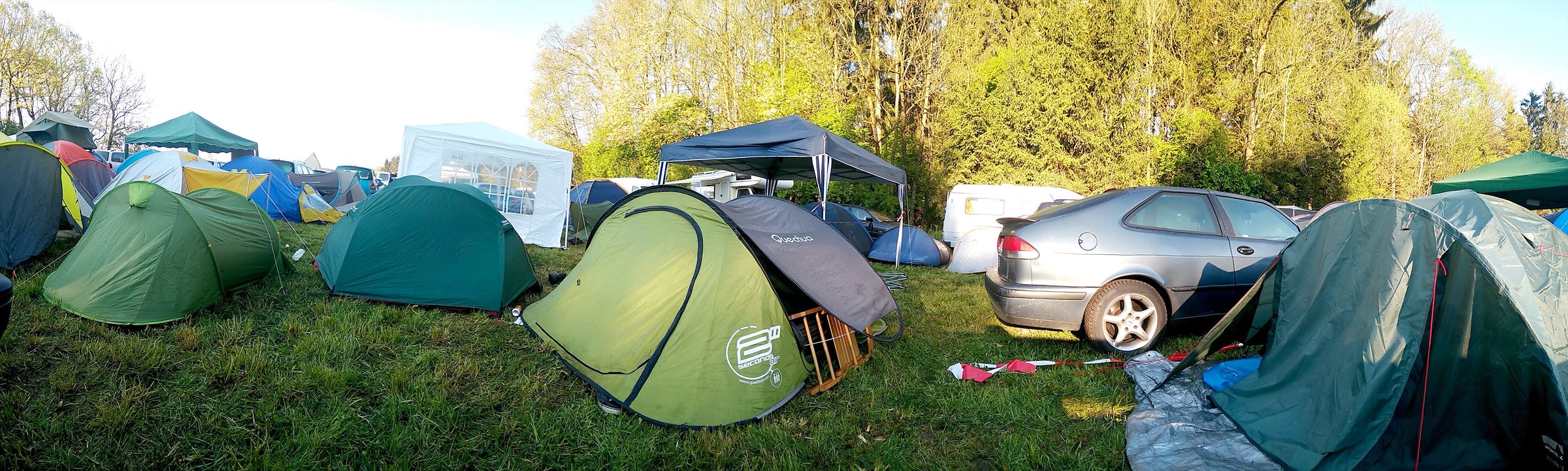 Zeltplatz am Morgen: Kuppelzelte, ein grünes Wurfzelt und ein Pavillon auf einer Wiese, dahinter Wohnmobile und Bäume im Gegenlicht.
